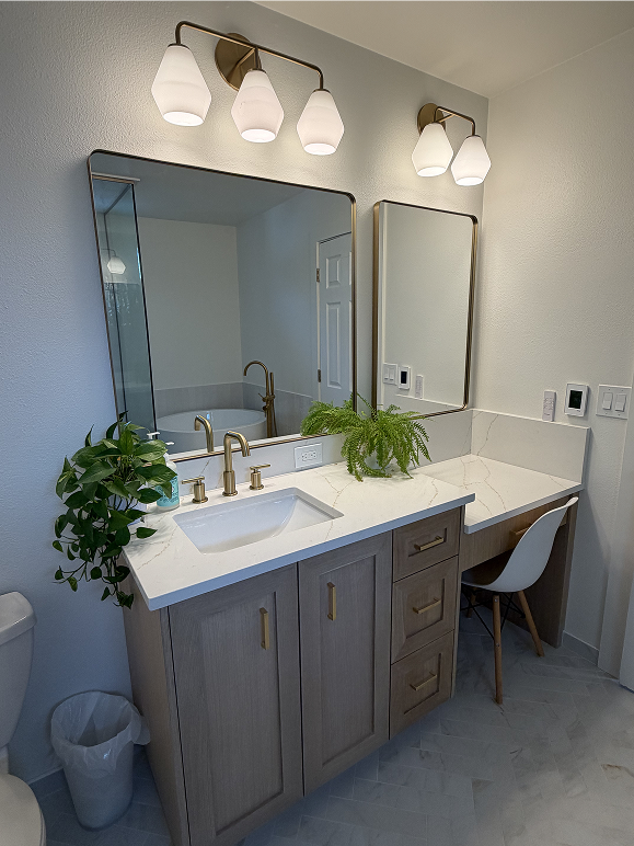 Light wood bathroom vanity with white quartz countertops, brass faucets, rectangular brass-framed mirrors, and a seated vanity desk.
